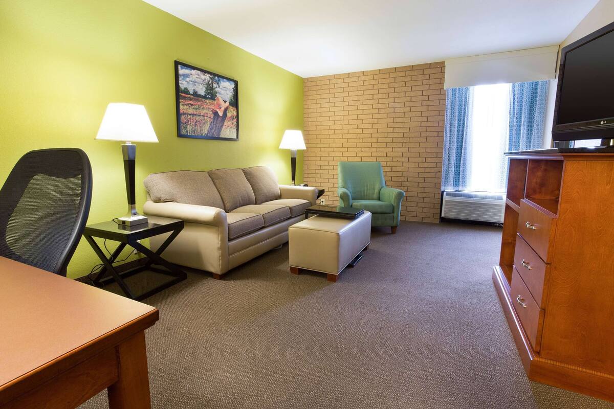 A hotel room with a green wall, a beige sofa, a small table, a green armchair, a wooden dresser, and a television.