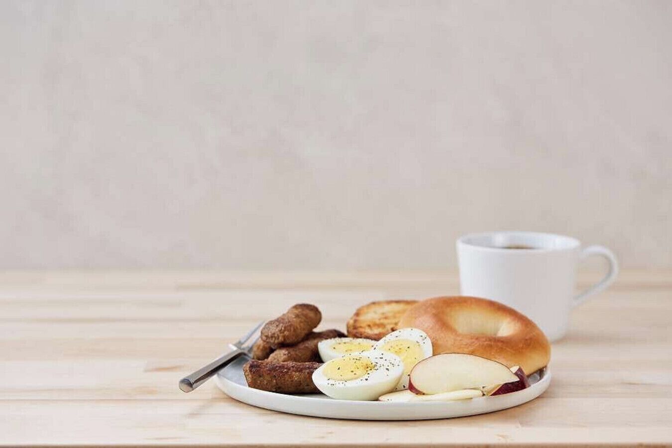 Prato de café da manhã com ovos, salsicha e um bagel, acompanhado de uma xícara de café sobre uma mesa de madeira.