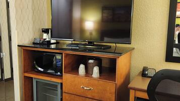 A compact hotel room with a wooden desk, a flat-screen TV, a microwave, and a small refrigerator.