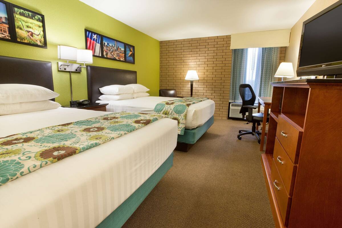 A hotel room with two beds, a desk, a chair, a TV, and a lamp.