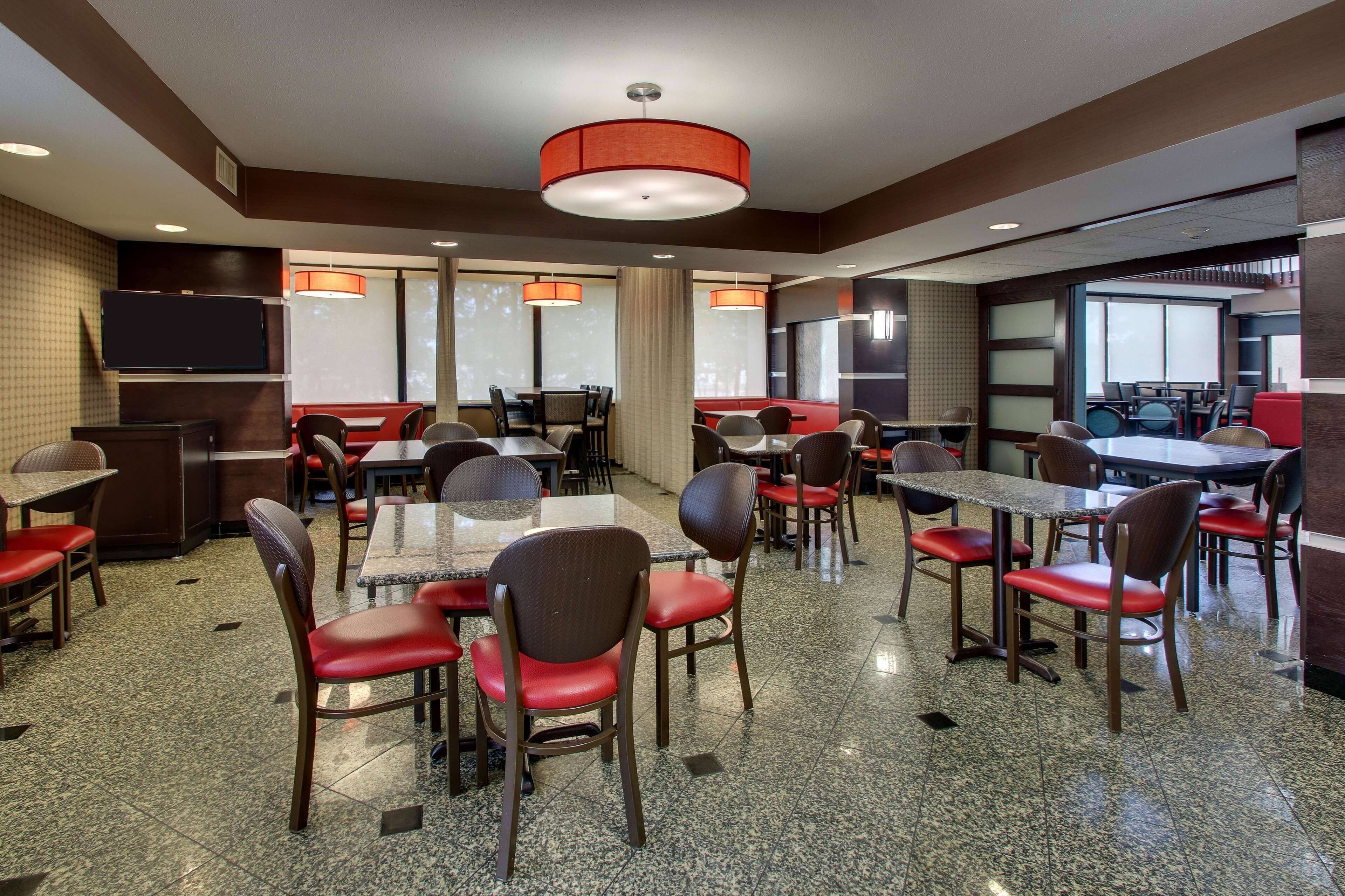 A hotel dining area with multiple tables and chairs, a television mounted on the wall, and large windows allowing natural light.