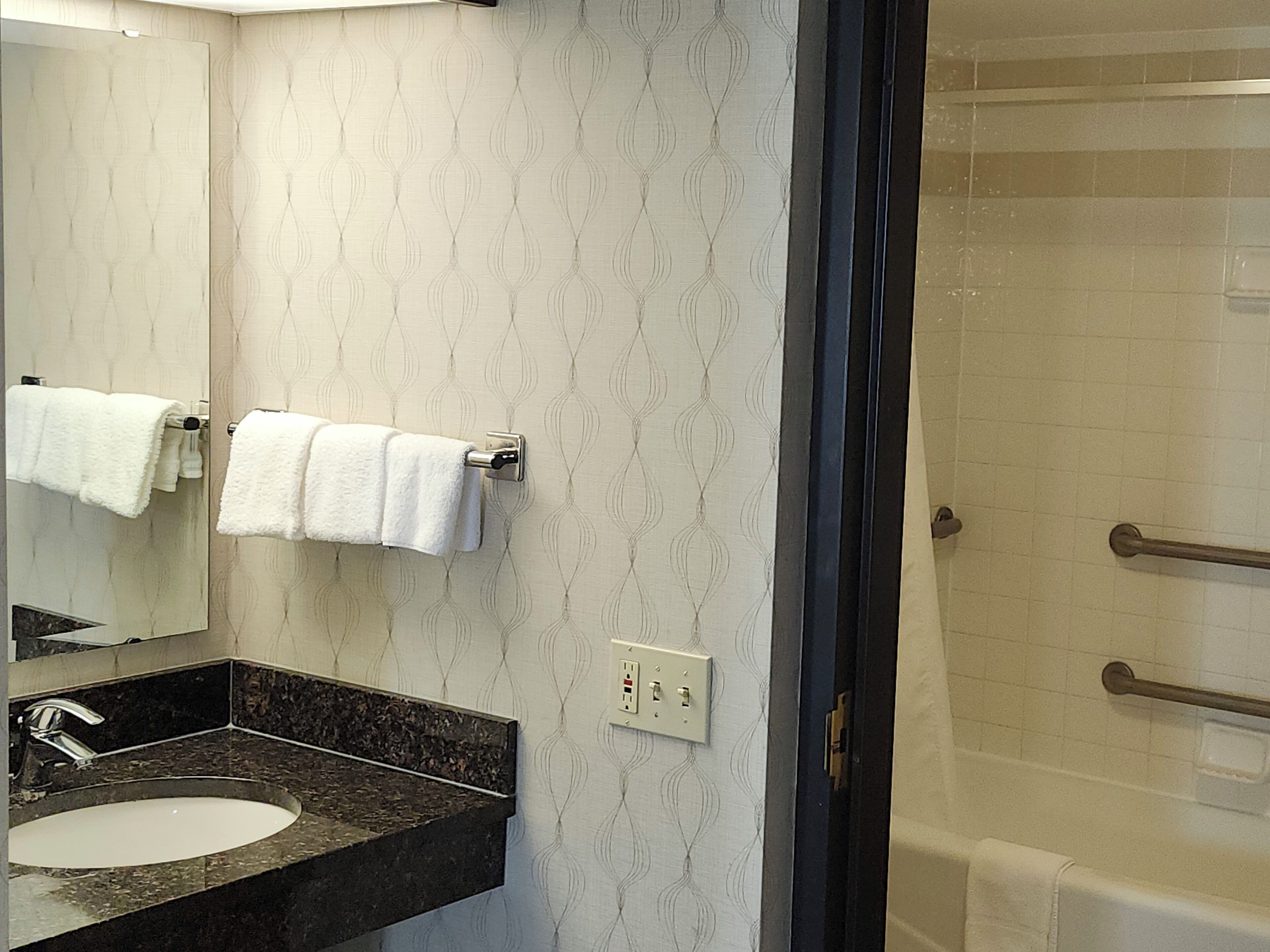 A bathroom with a marble sink, a mirror, two towels on towel racks, a shower with a glass door, and a wall with a subtle pattern.