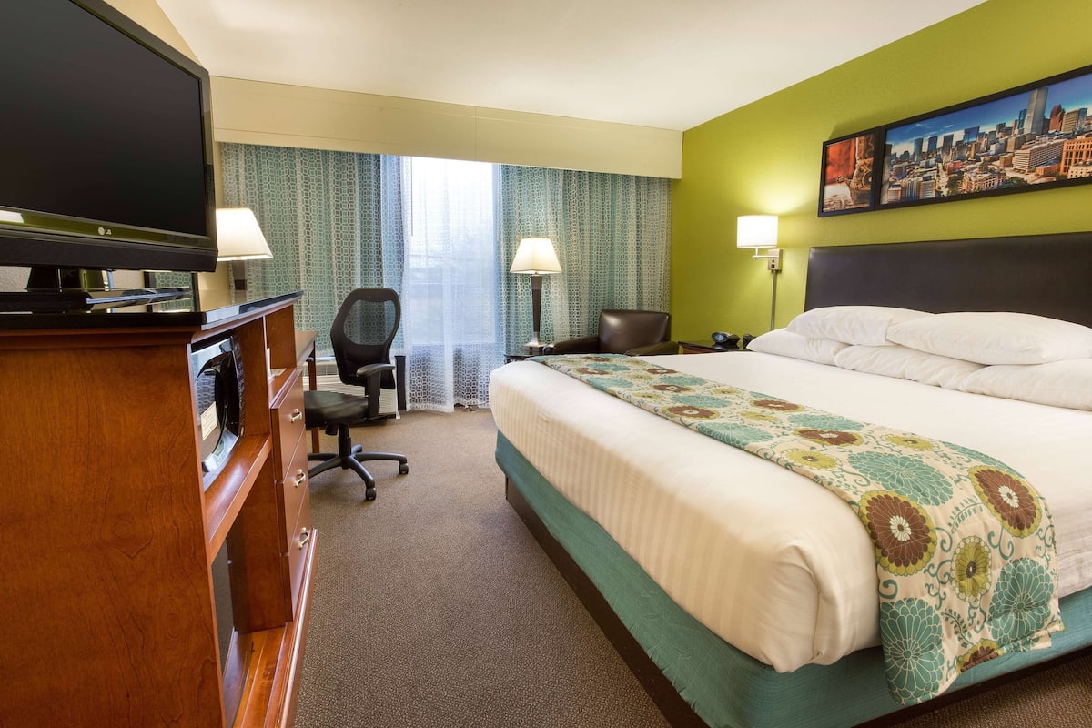 A hotel room with a large bed, a desk with a flat-screen TV, two chairs, a lamp, and a painting on the wall.