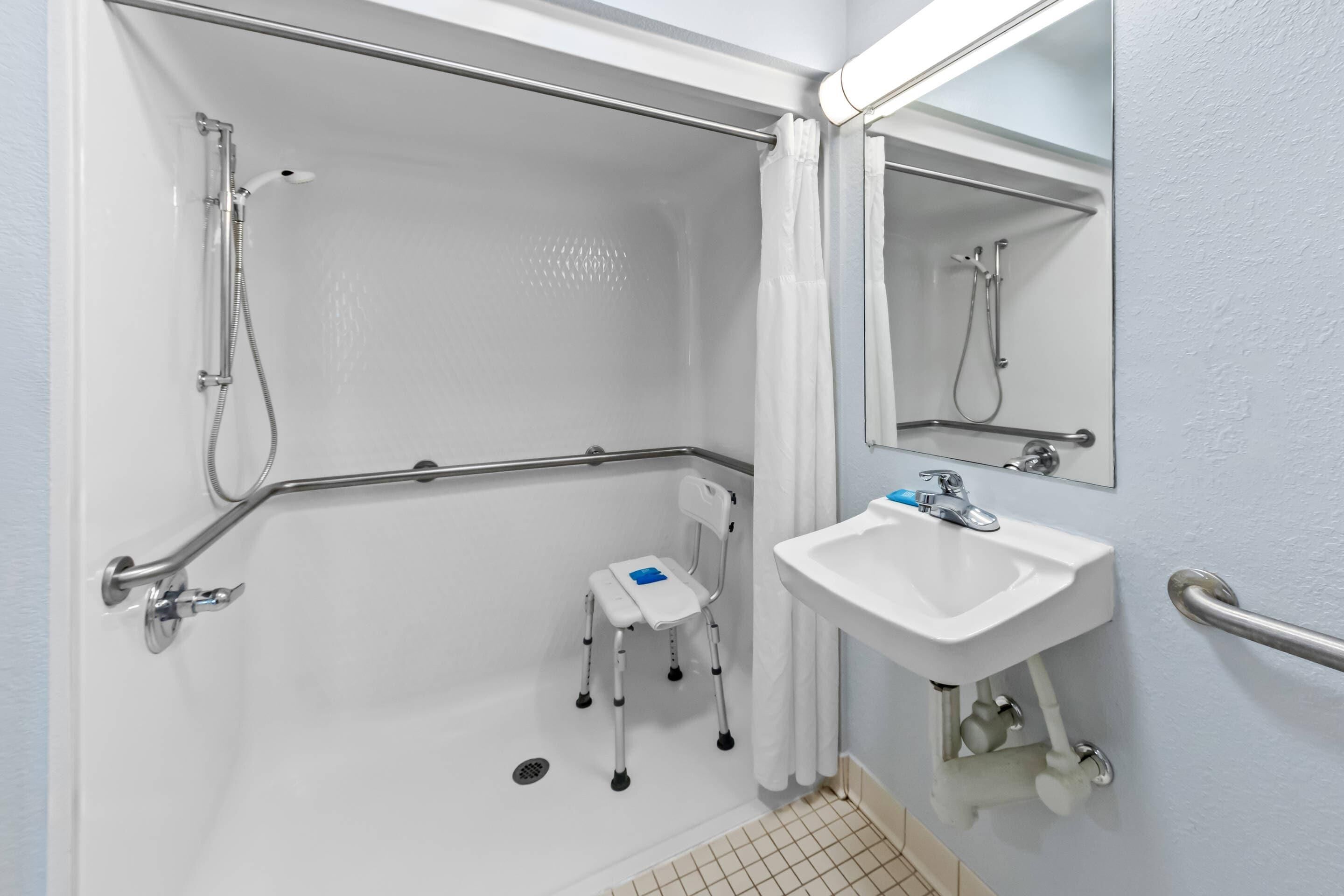 A bathroom with a shower, a sink, a mirror, and a wheelchair-accessible bench.