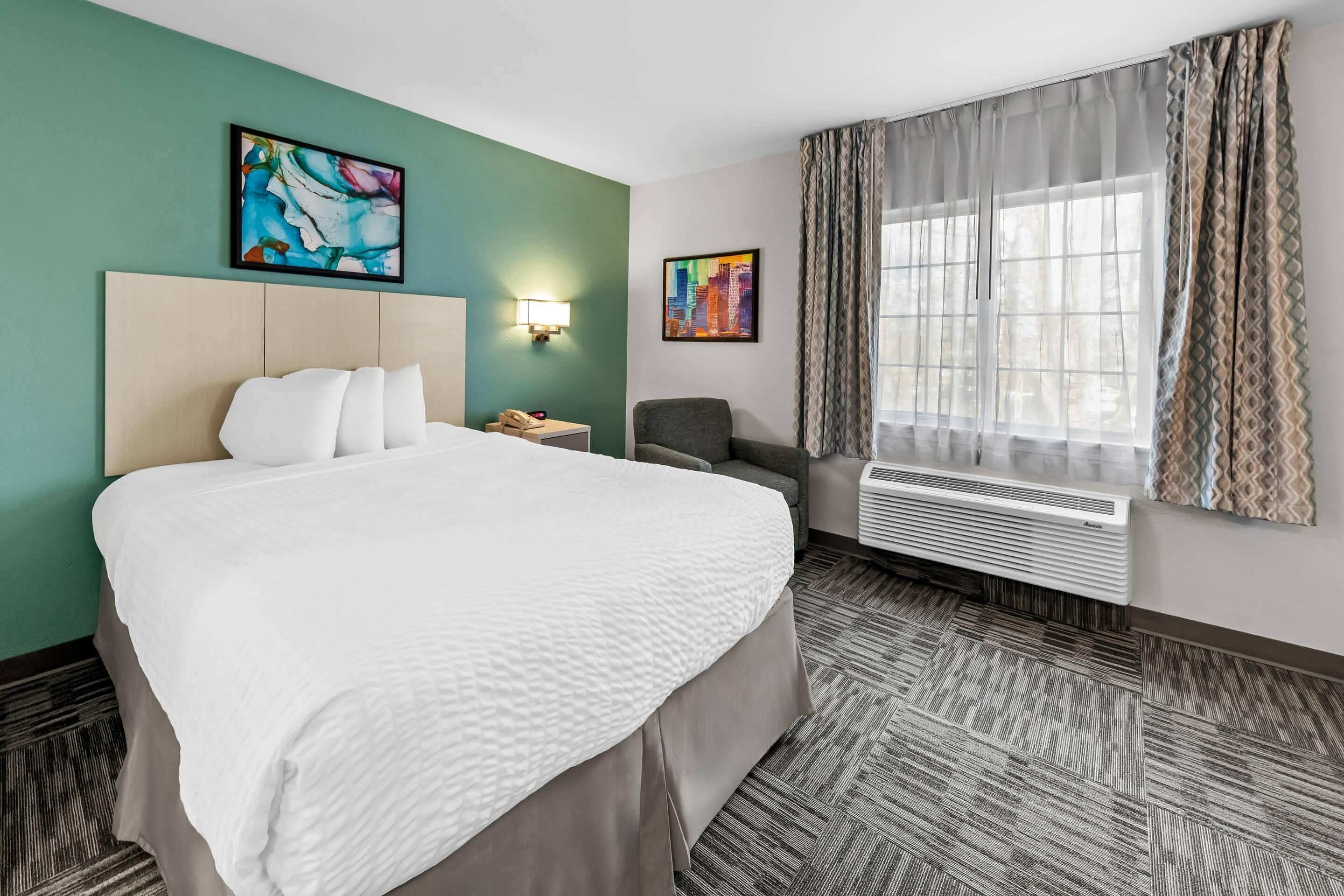 A hotel room with a large bed, a desk with a lamp, a chair, a window with curtains, and two paintings on the walls.