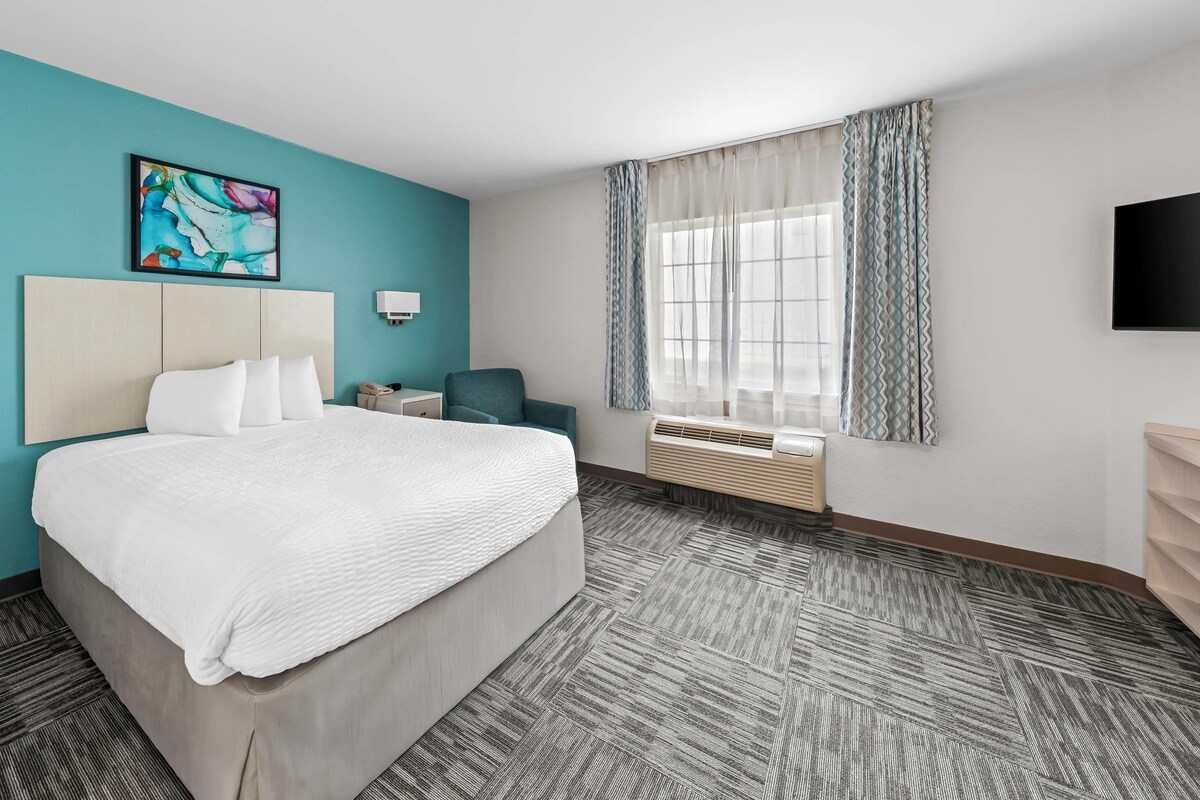 A hotel room with a bed, a desk, a chair, a window with curtains, a flat-screen TV, and a wall-mounted air conditioner.