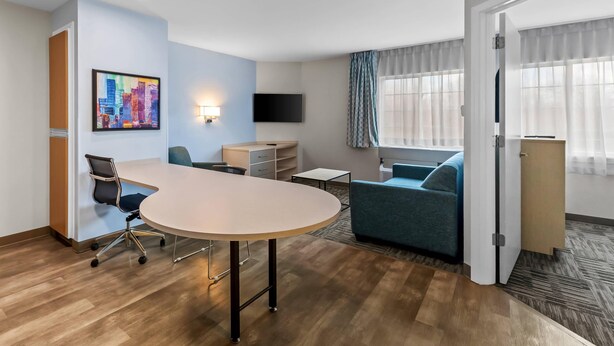 A hotel room with a round table, a sofa, a TV, and a cityscape painting. - MainStay Suites Cincinnati Blue Ash (Cincinnati)