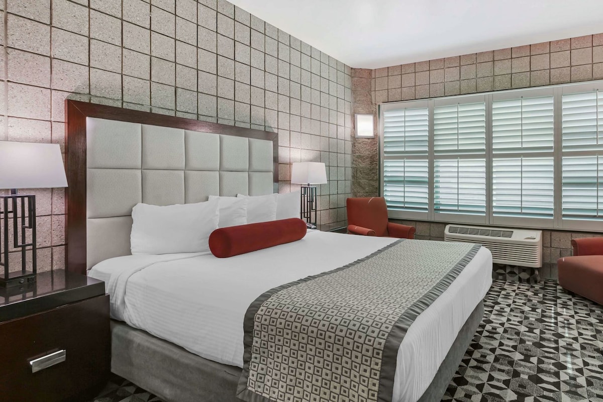 A hotel room with a large bed, a headboard, a desk with a lamp, a chair, and a window with blinds.