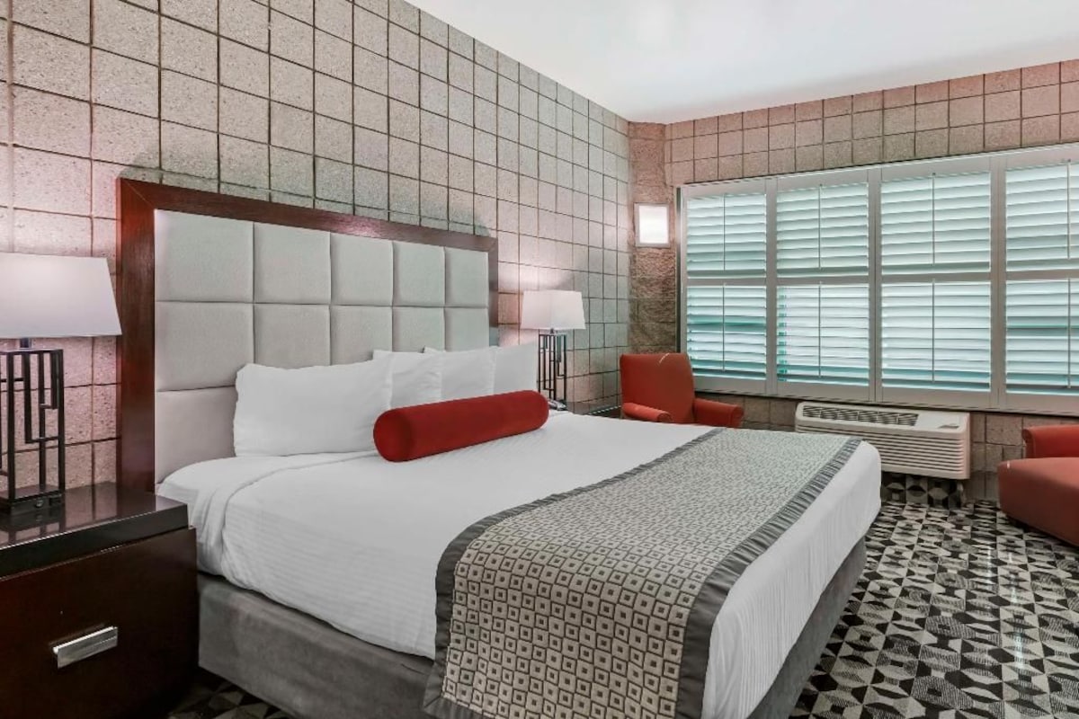 A hotel room with a large bed, a nightstand, a chair, a window with blinds, and a patterned floor.