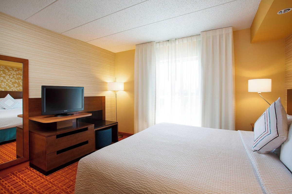 A hotel room with a bed, a TV on a wooden stand, two bedside lamps, and a window with curtains.