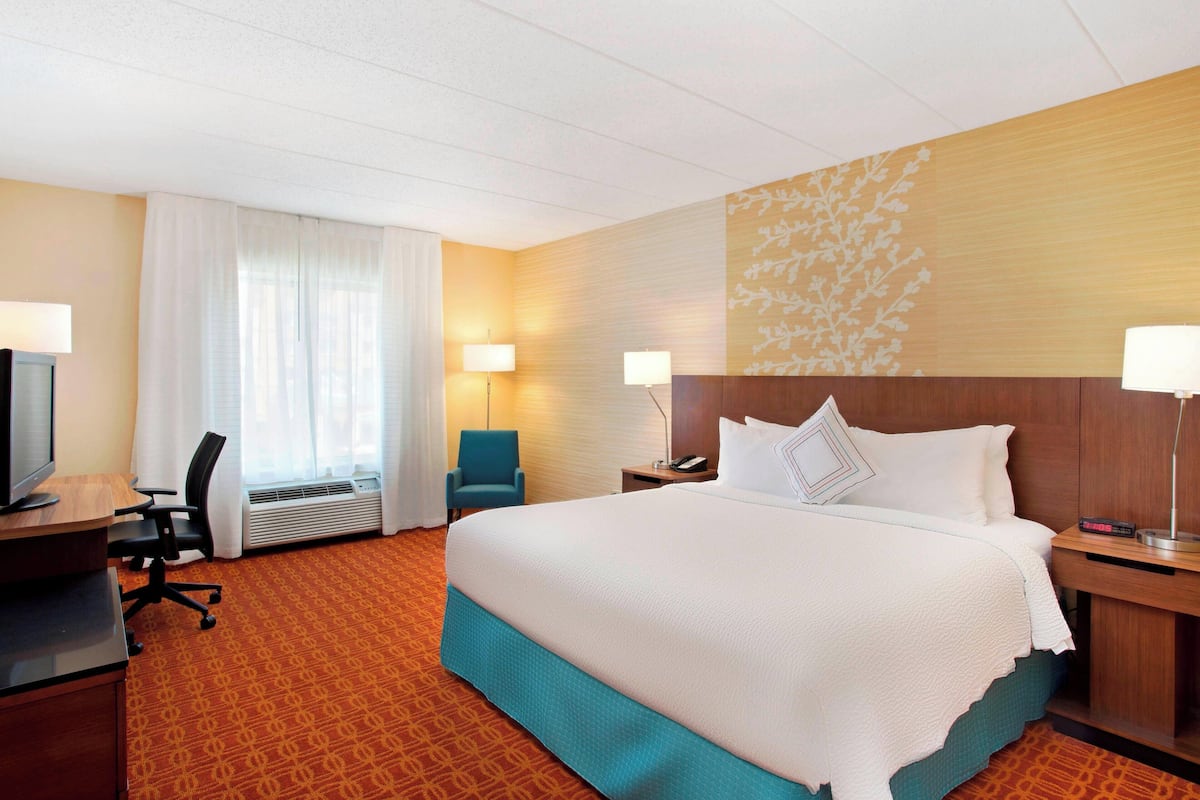 A hotel room with a large bed, a desk with a computer, a chair, a desk lamp, a bedside table with a lamp, and a wall with a floral pattern.