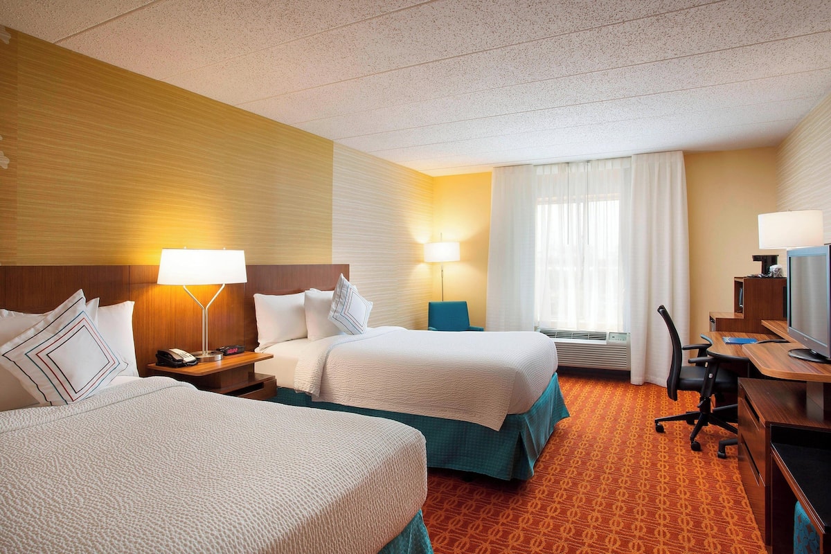 A hotel room with two beds, a desk, a chair, a TV, and a window with curtains.
