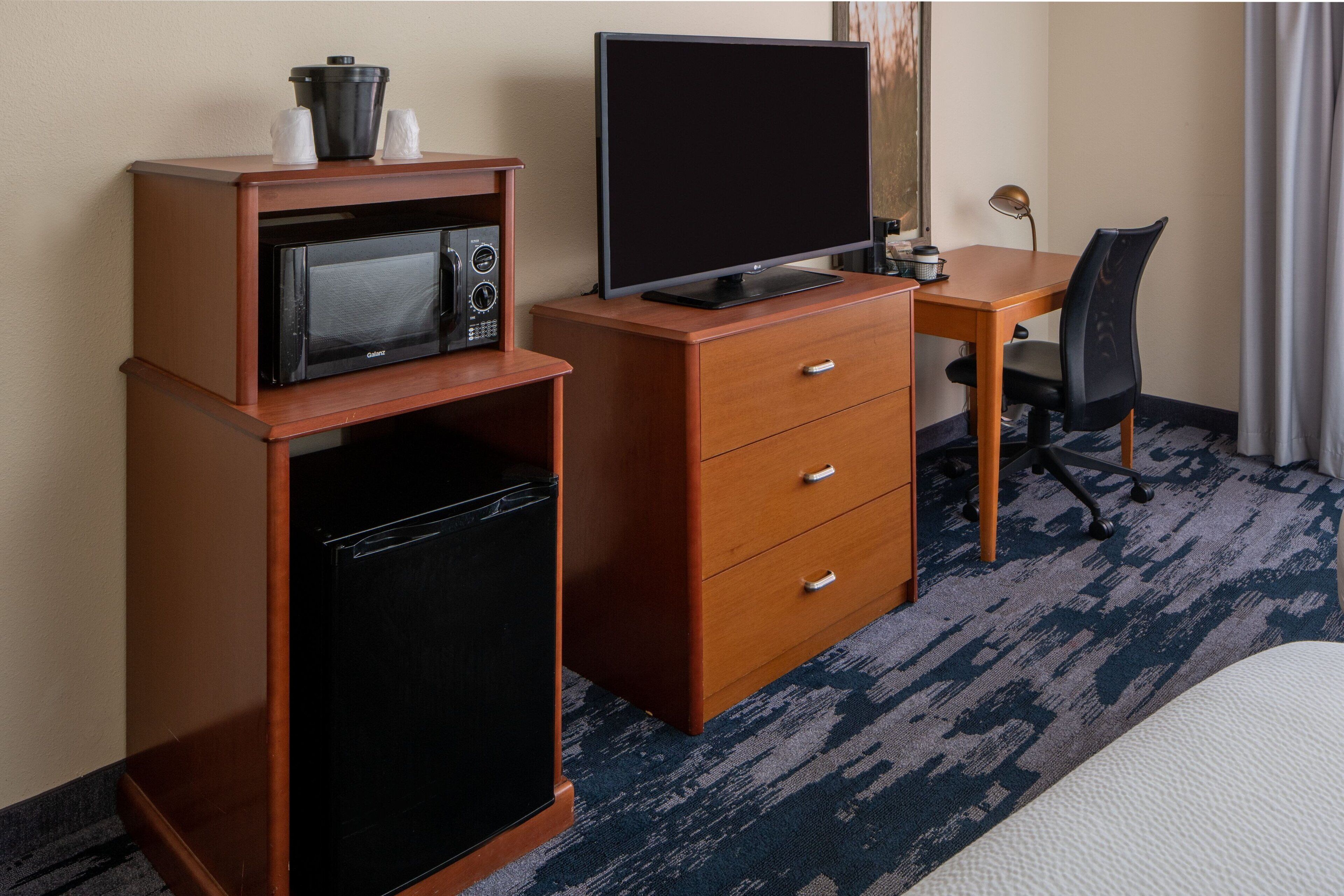 A compact hotel room with a kitchenette, including a microwave, refrigerator, and a small TV on a wooden cabinet. There's a desk with a chair and a bed with a white comforter.