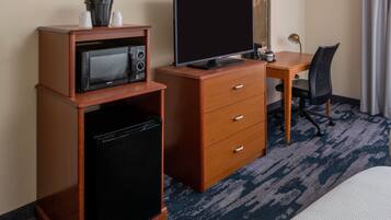 A compact hotel room with a kitchenette, including a microwave, refrigerator, and a small TV on a wooden cabinet. There's a desk with a chair and a bed with a white comforter.