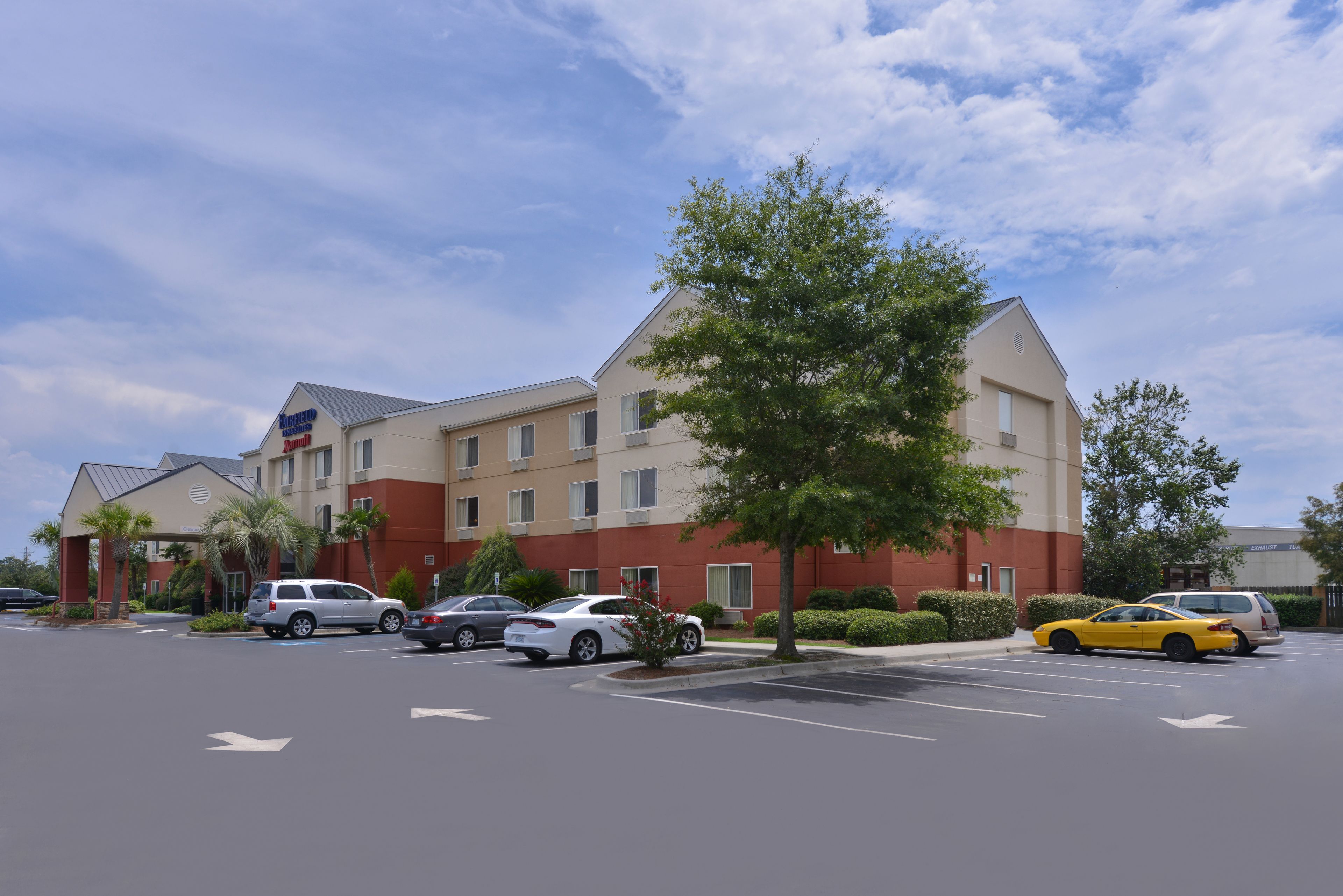 A hotel with a parking lot, several cars, and a large tree in front.