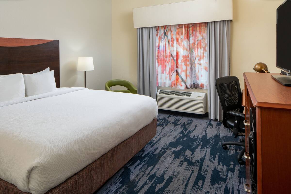 A hotel room with a large bed, a desk with a computer, a chair, an air conditioning unit, and a window with curtains.
