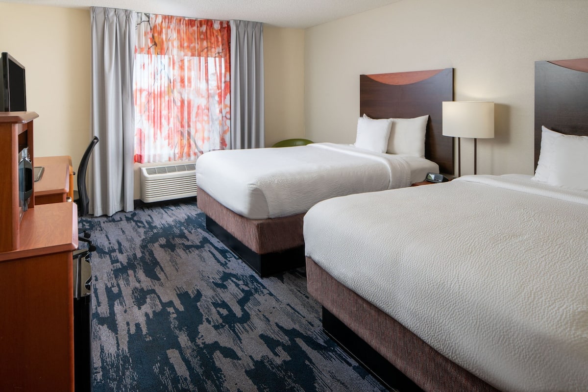 A hotel room with two beds, a desk, a chair, a TV, and a window with curtains.