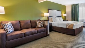 Una habitació d'hotel amb un sofà marró, un llit amb roba de llit blanca i una paret verda decorativa.