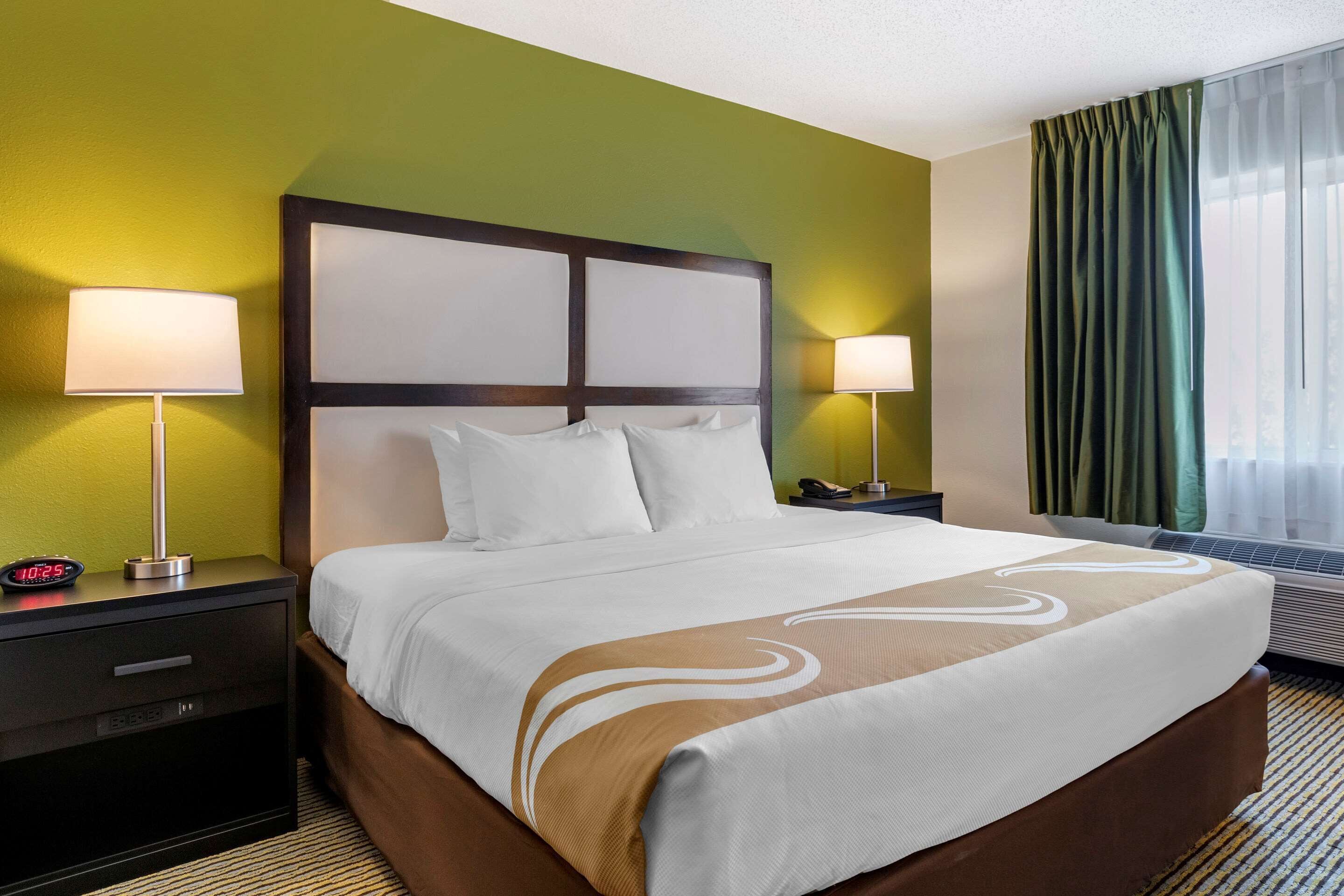A hotel room with a large bed, two bedside tables, a nightstand, a wall clock, and a window with curtains.