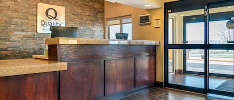 Un área de recepción con una pared de piedra, un mostrador de madera y un reloj digital que muestra las 10:38.