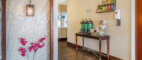 A hallway with a wooden floor, a decorative wall with geometric patterns, a hanging lantern, a table with coffee dispensers, and a shelf with books and a coffee machine.