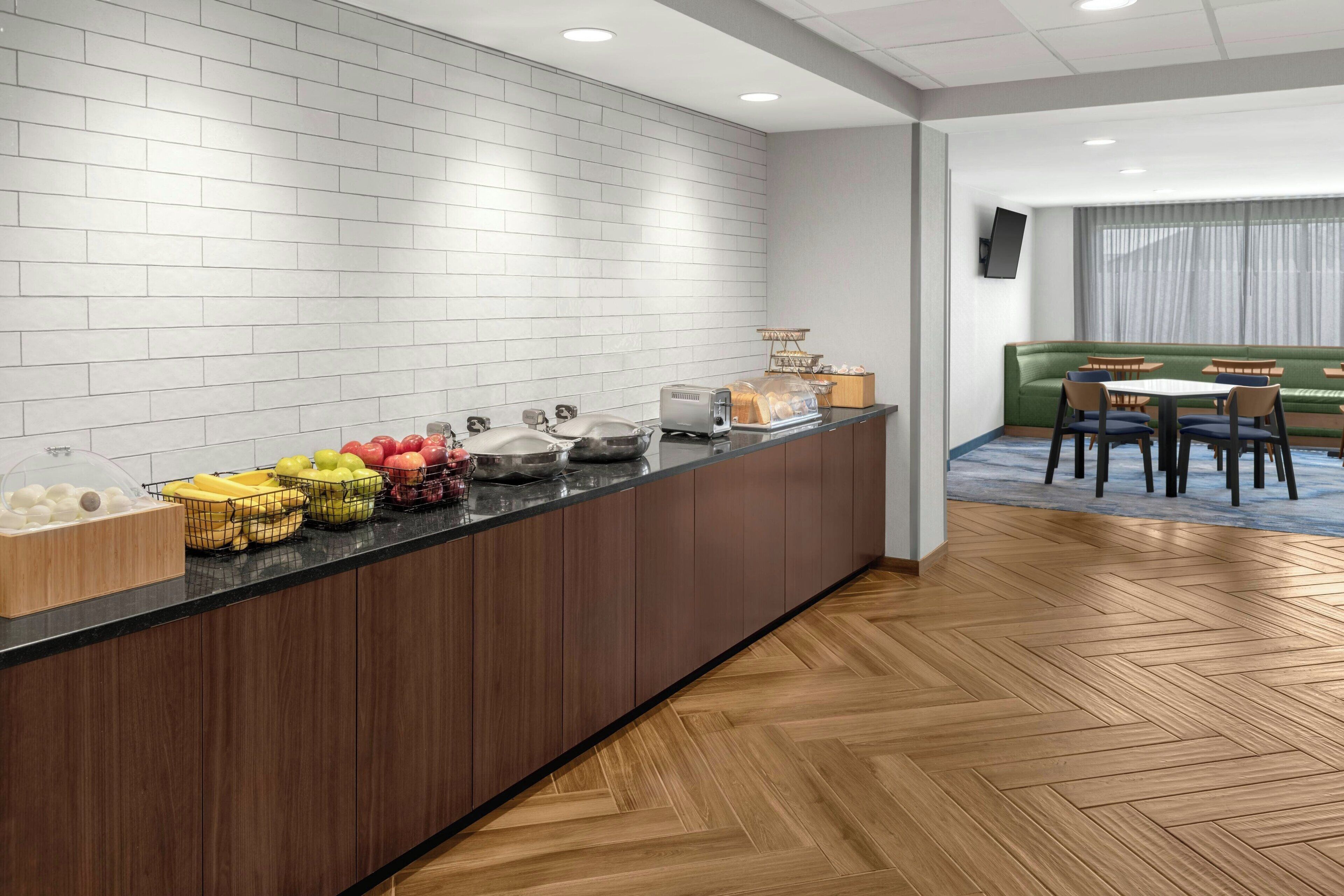 A modern kitchen area with a long counter, various fruits, a coffee machine, and a seating area with tables and chairs.