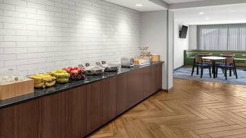 A modern kitchen area with a long counter, various fruits, a coffee machine, and a seating area with tables and chairs.