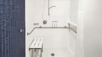Área de duche com chão em azulejo branco, chuveiro, mangueira de duche e um pequeno banco.