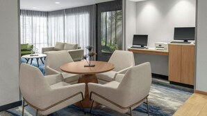 Una habitación de hotel moderna con una mesa de comedor redonda, cuatro sillas, un sofá, un escritorio con dos monitores y un ventanal con vistas a un bosque.