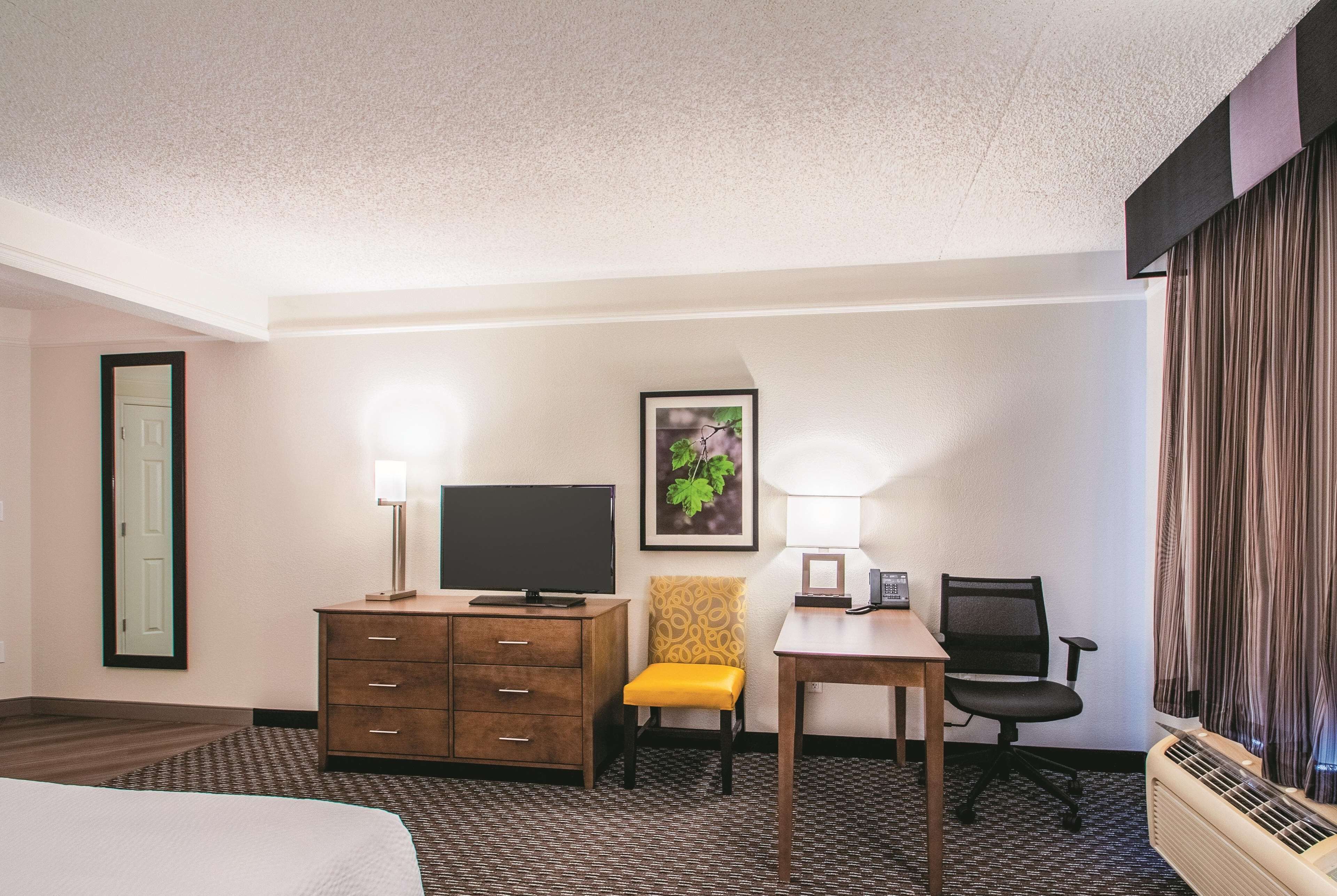 A hotel room with a bed, a desk with a chair, a television, a lamp, a framed picture, and a window with curtains.