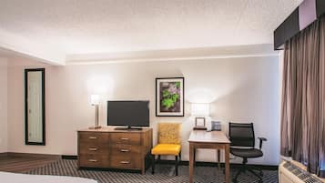A hotel room with a bed, a desk with a chair, a television, a lamp, a framed picture, and a window with curtains.