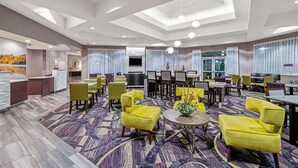 A hotel lobby with a central seating area, multiple tables, and a large TV screen.