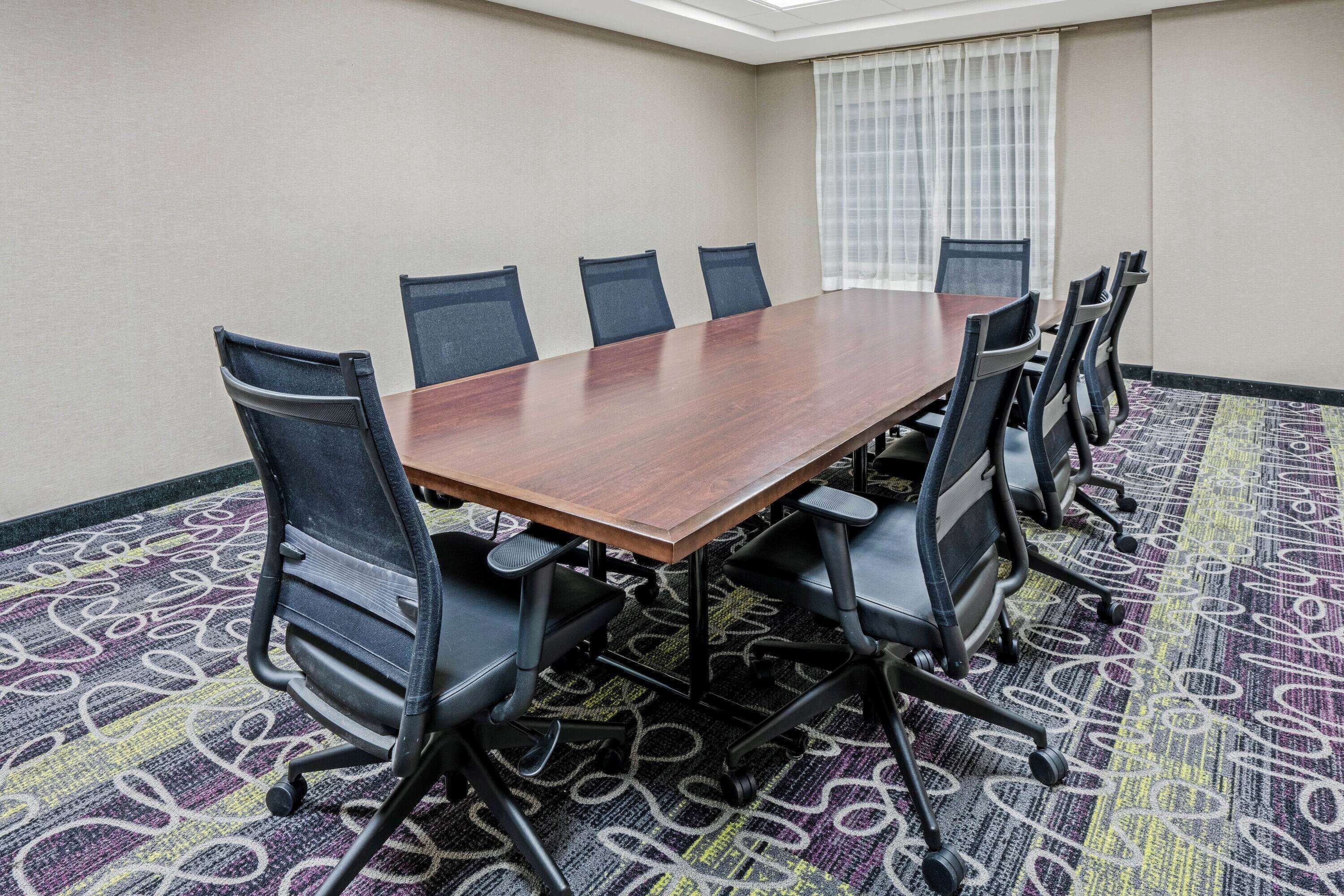 Une salle de conférence avec une longue table en bois, six chaises noires et un tapis à motifs.