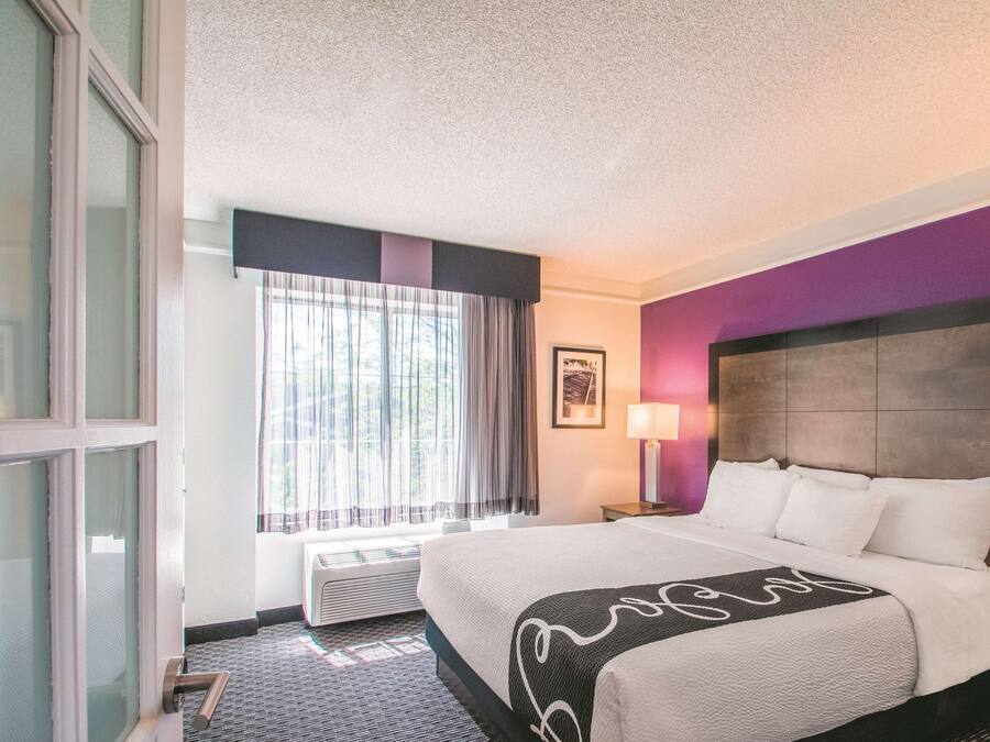 A hotel room with a large bed, a window with curtains, a framed picture on the wall, and a bedside lamp.