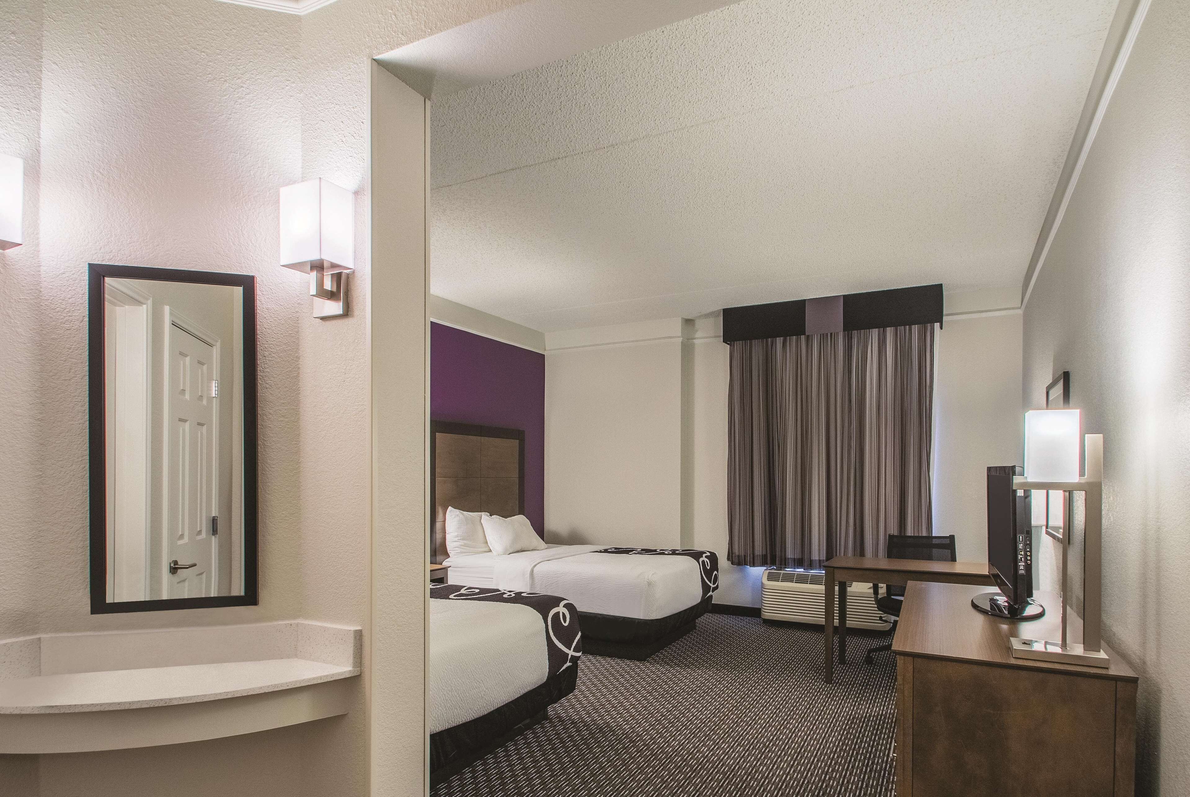 A hotel room with two beds, a desk, a TV, and a bathroom with a sink and mirror.