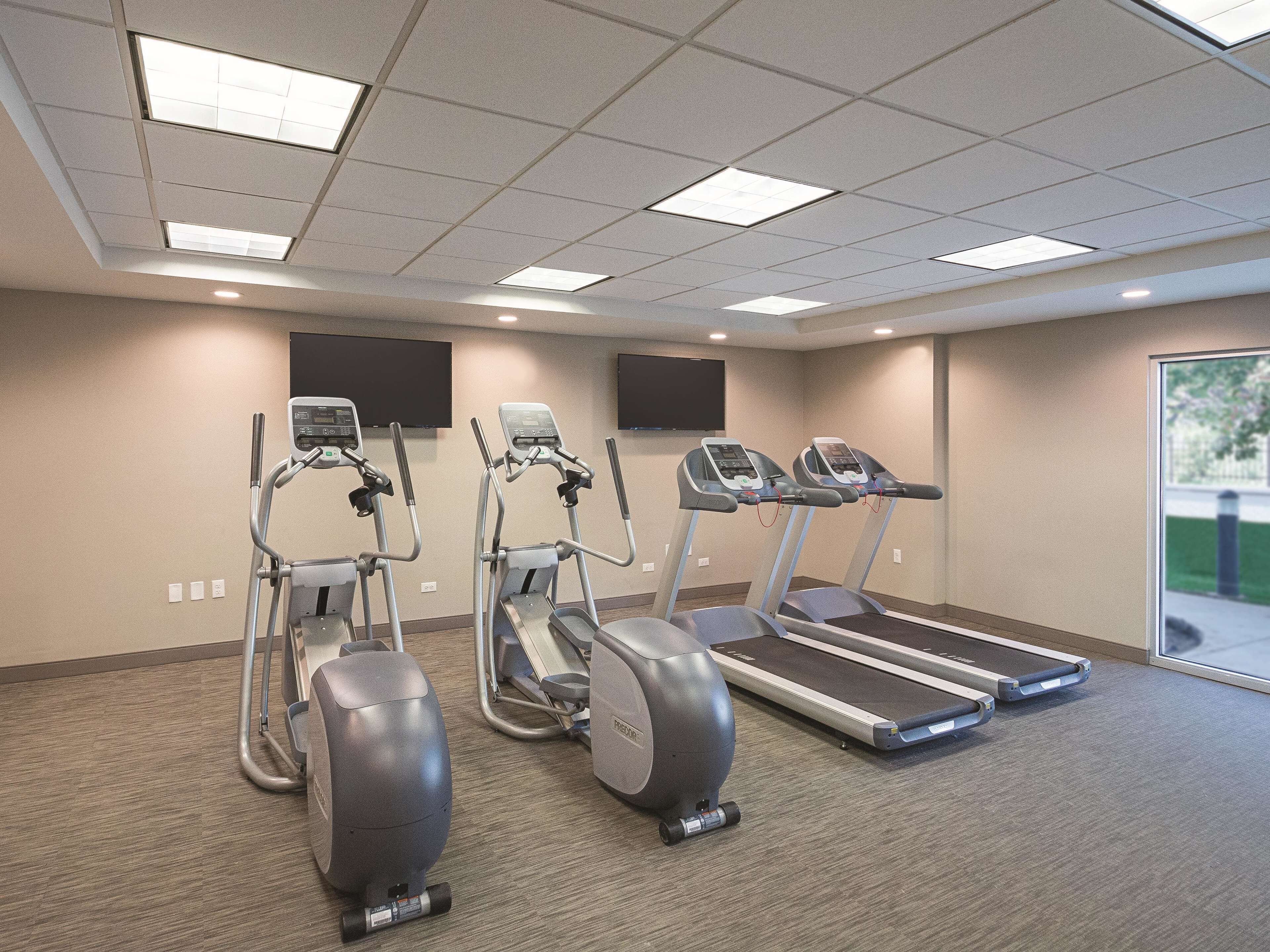 Een fitnessruimte met loopbanden en elliptische trainers, een vloerbedekking en een groot raam met uitzicht op de omgeving.