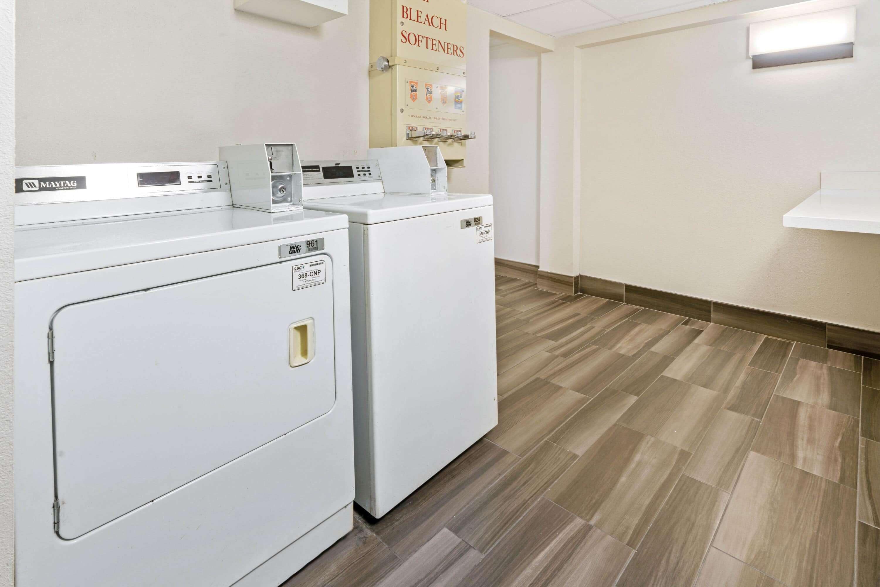 A laundry area with two Maytag washers and dryers, a sign for bleach softeners, and a light fixture on the wall.