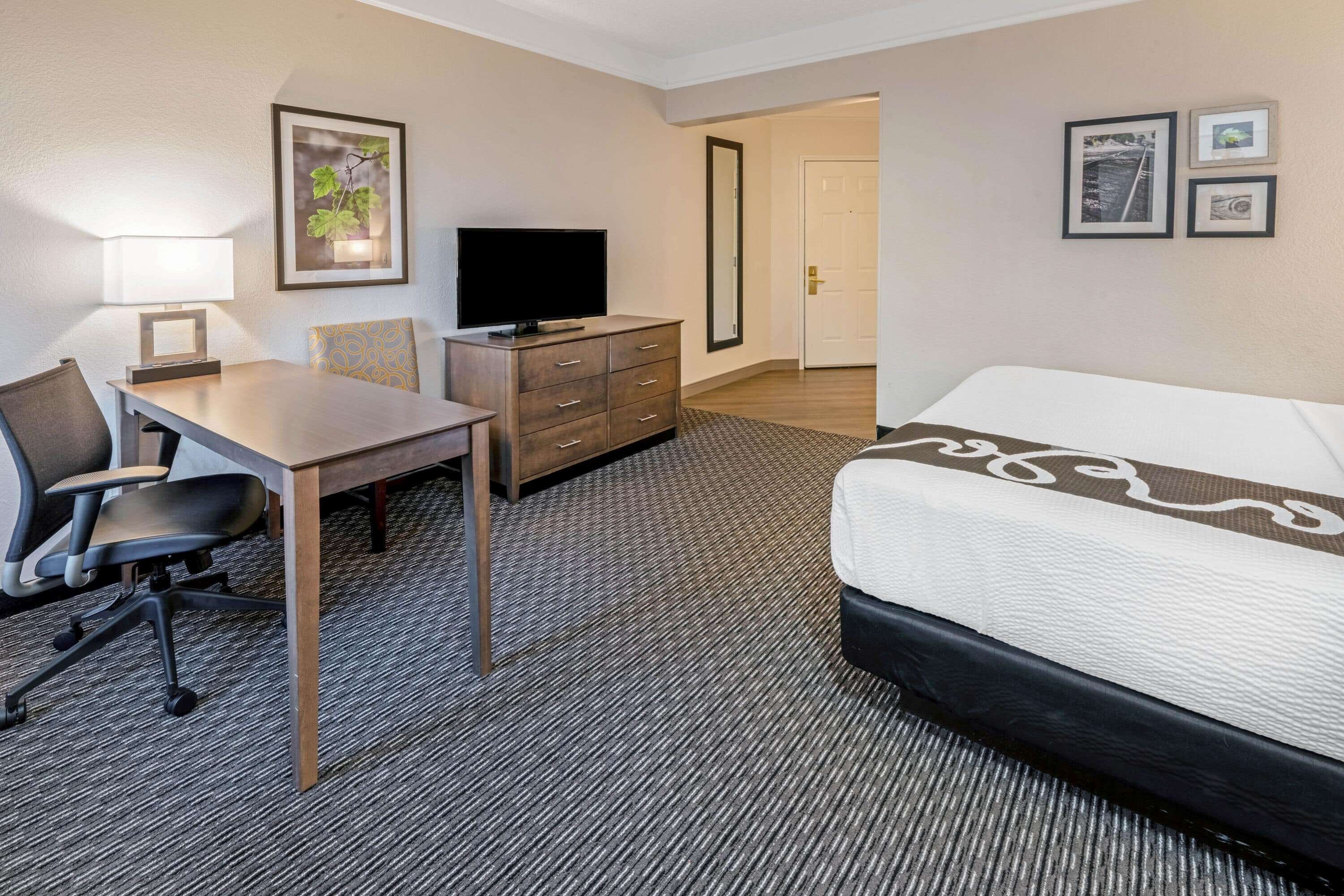 A hotel room with a bed, desk, chair, television, dresser, and framed pictures on the wall.