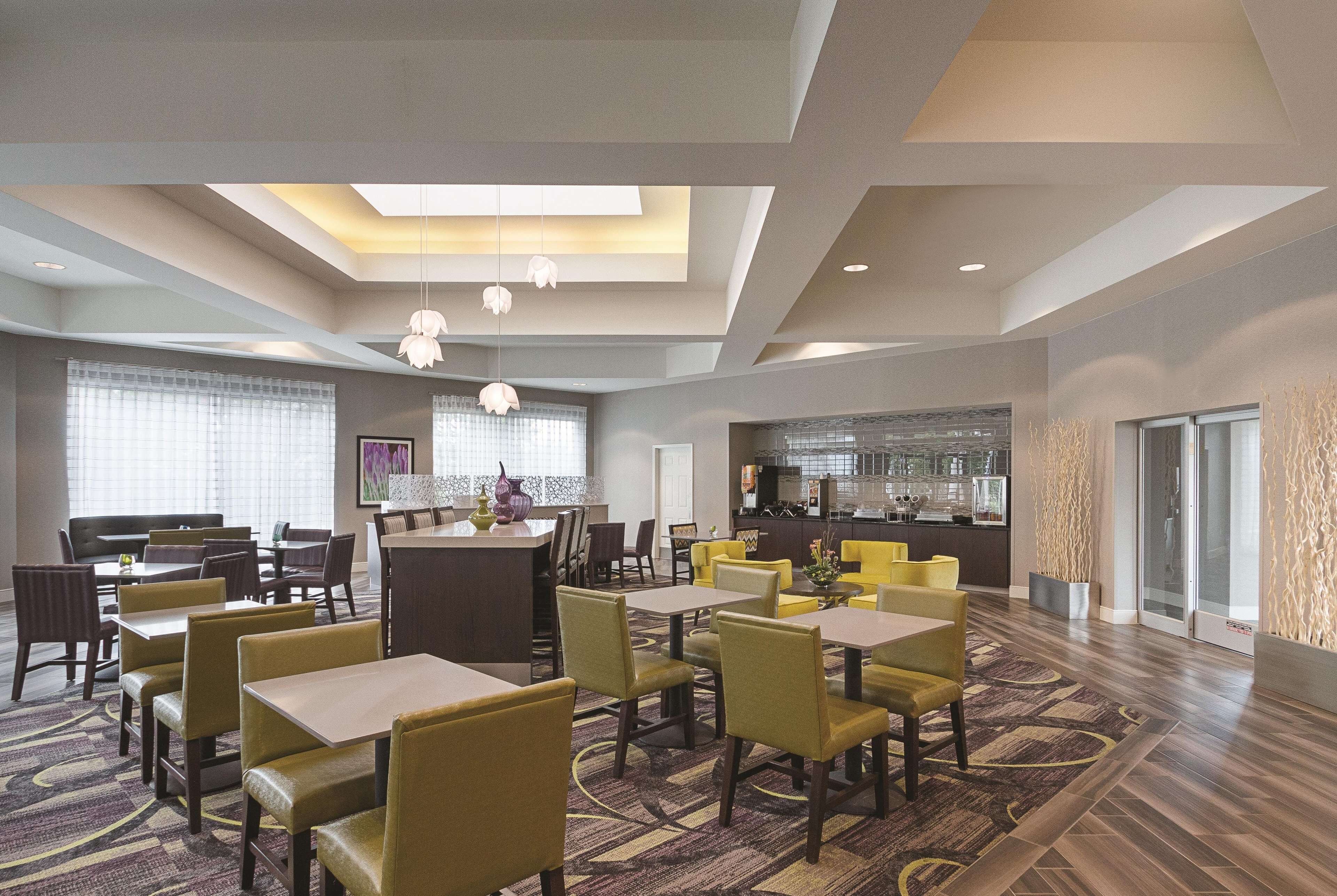 A hotel dining area with multiple tables and chairs, a kitchen in the background, and a large window with blinds.