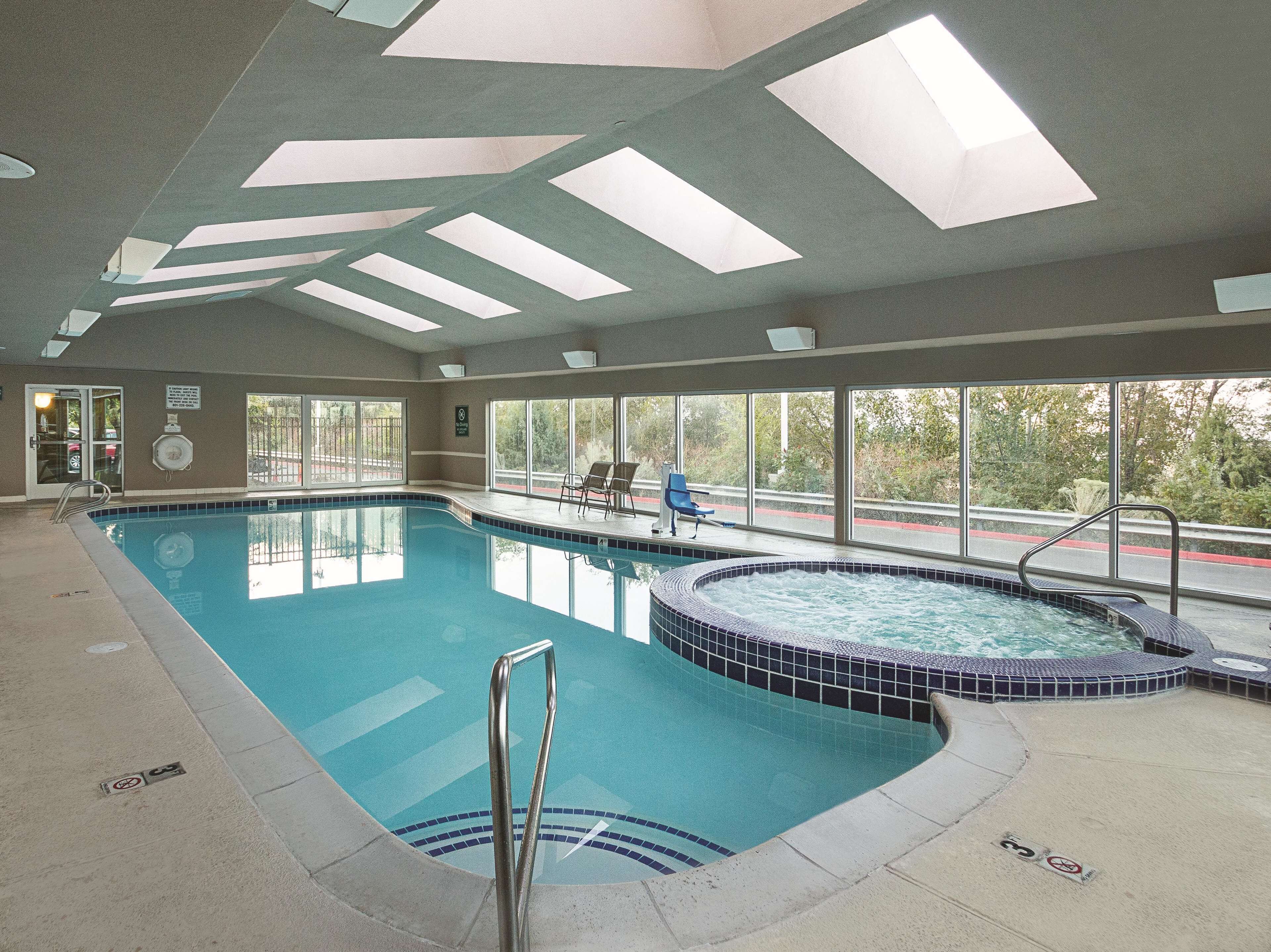 An indoor swimming pool with a hot tub, clear glass doors, and large windows offering a view of the outdoors.