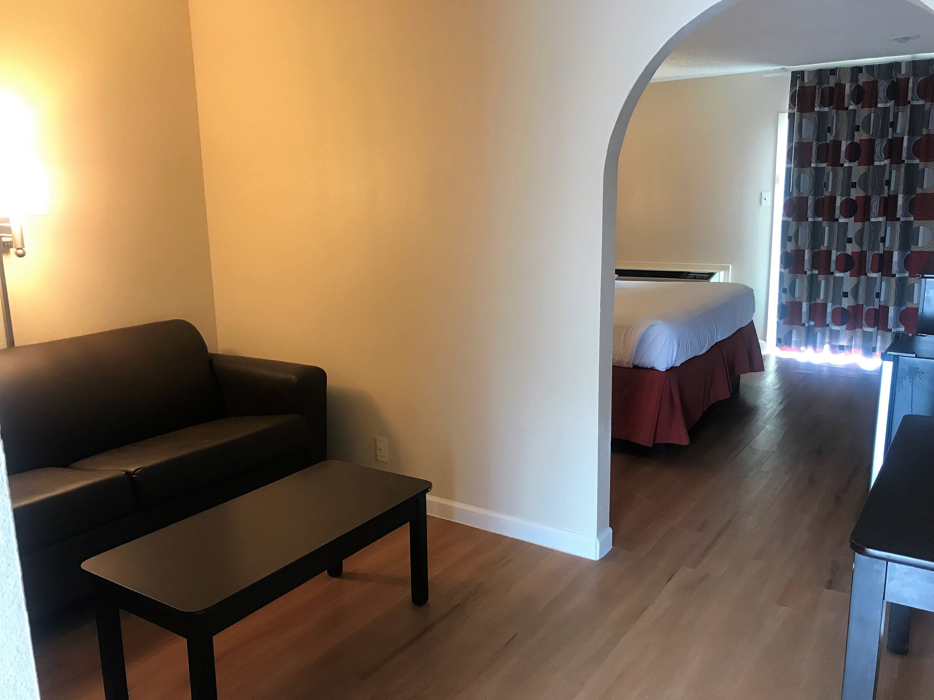 A room with a sofa, a small table, a bed with a red bedspread, and a patterned curtain.