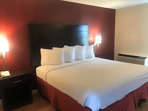A hotel room with a large bed, a nightstand, a lamp, and a wall-mounted heater. - Red Roof Inn & Suites San Angelo (San Angelo)