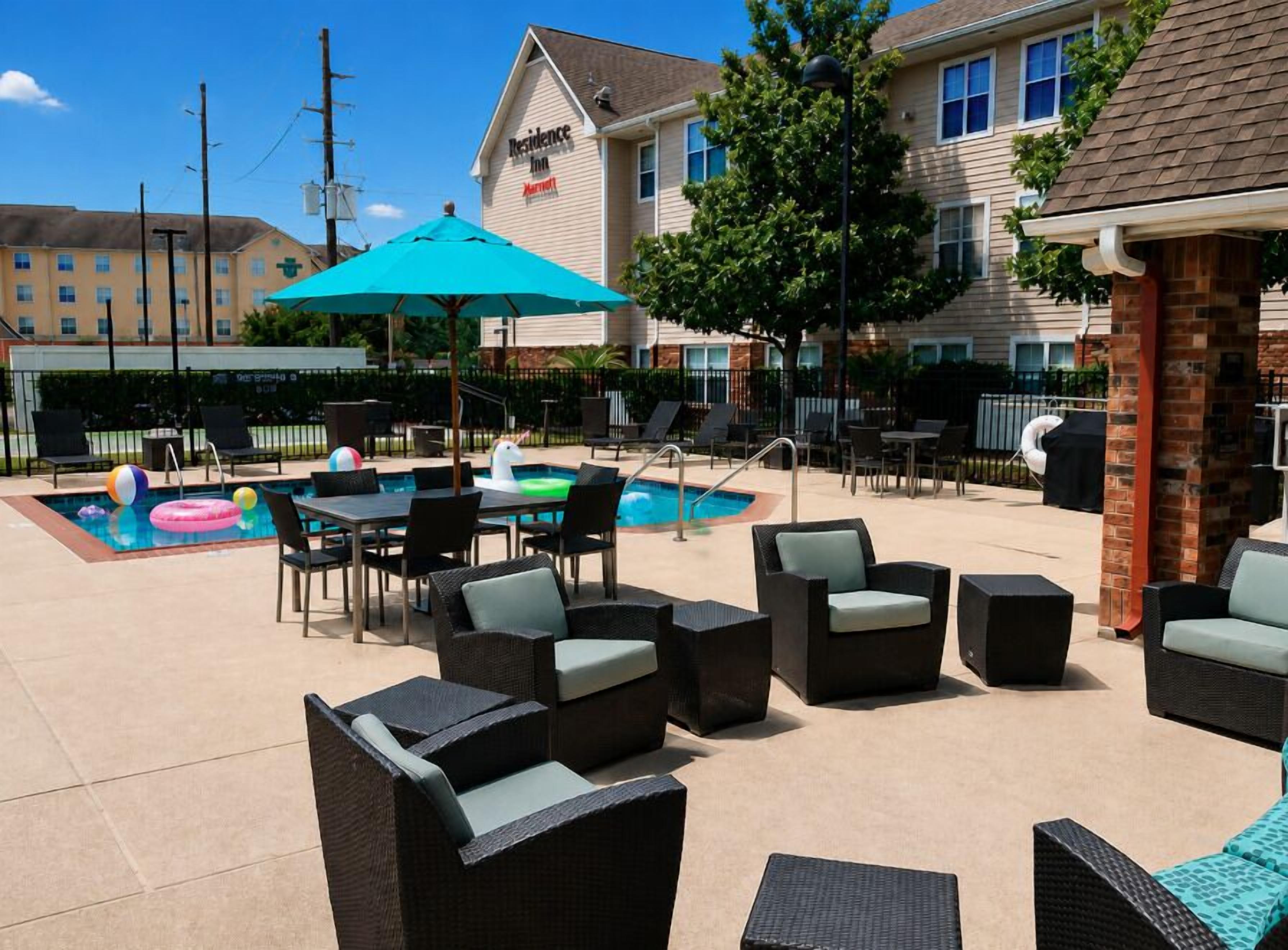 An outdoor pool area with lounge chairs and tables, a brick wall, and a building in the background.