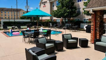An outdoor pool area with lounge chairs and tables, a brick wall, and a building in the background.