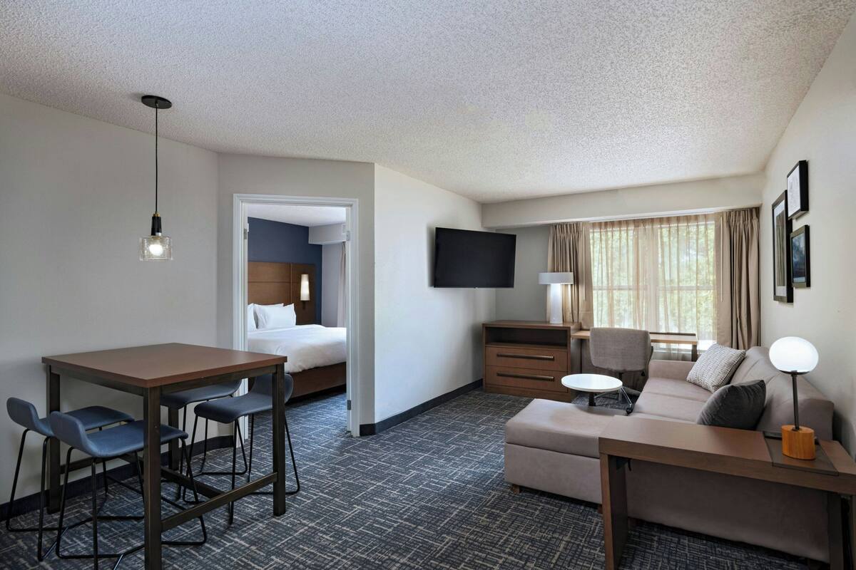 A hotel room with a bed, a desk with chairs, a sofa, a TV, and a window with curtains.