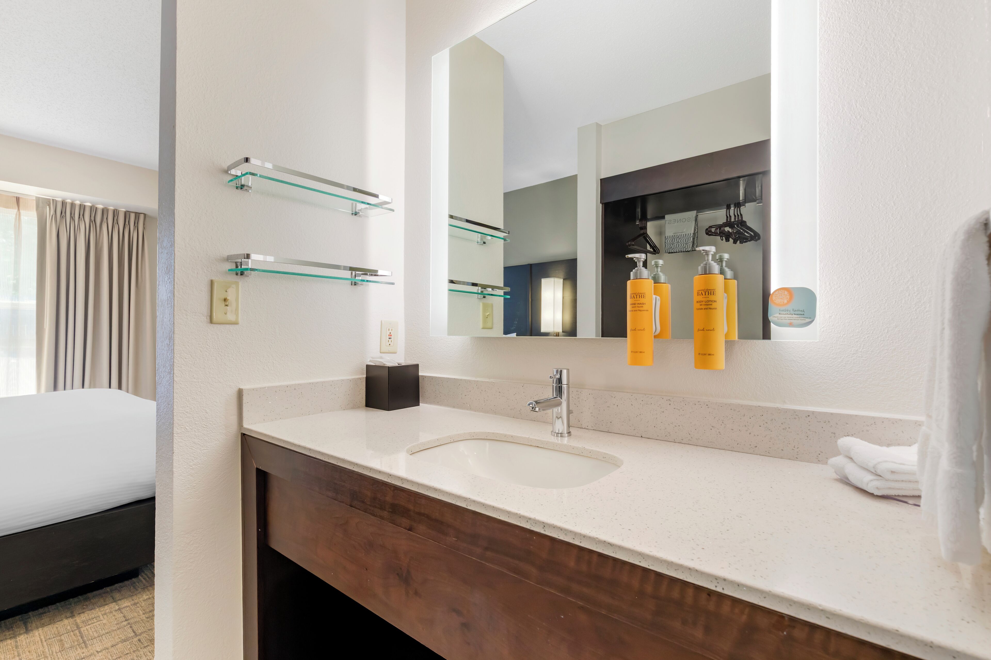 Een moderne badkamer met een wit wastafelmeubel, een grote spiegel en glazen planken met toiletartikelen.