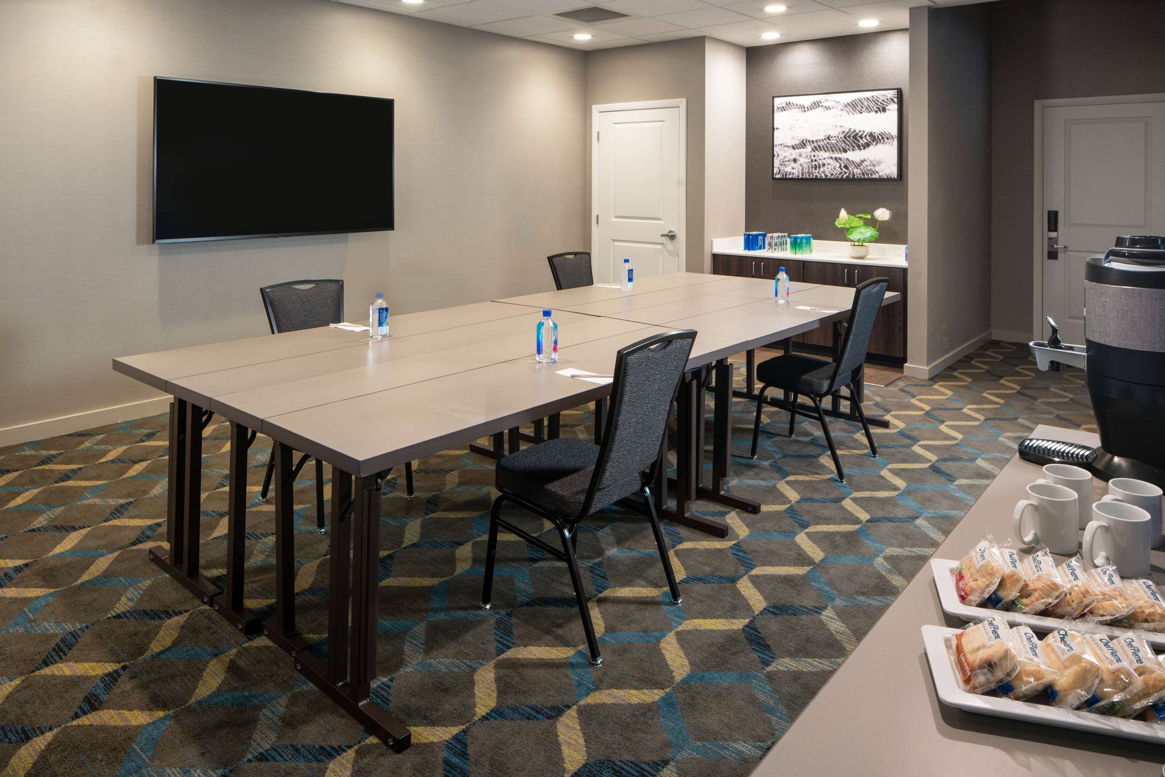 A conference room with a long table, chairs, a TV screen, a door, and a counter with water bottles and snacks.