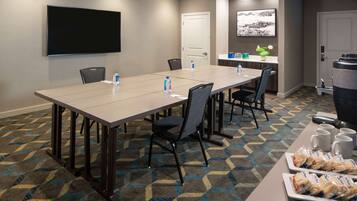 A conference room with a long table, chairs, a TV screen, a door, and a counter with water bottles and snacks.
