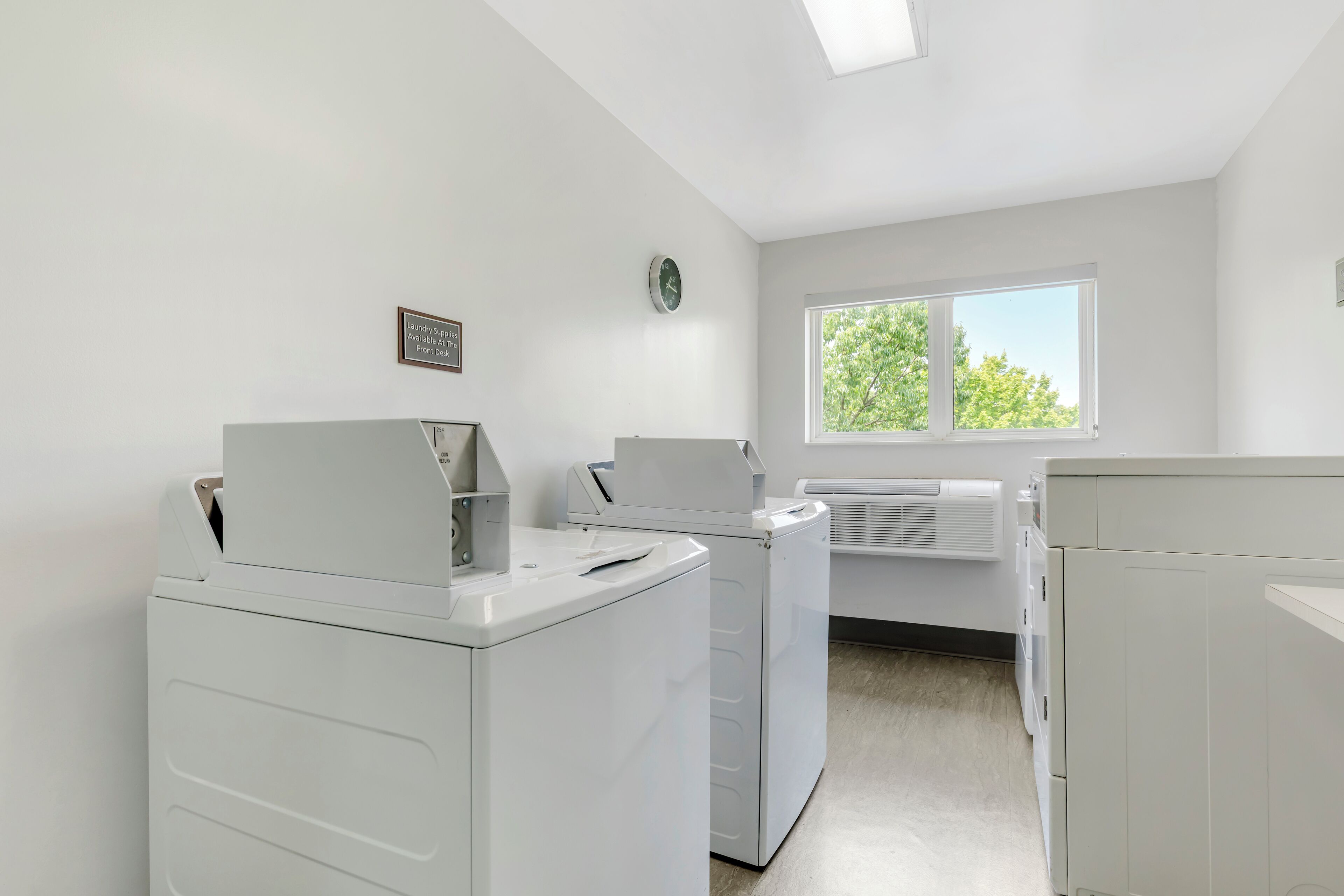 A laundry room with white appliances, a clock, and a window with a view of trees.