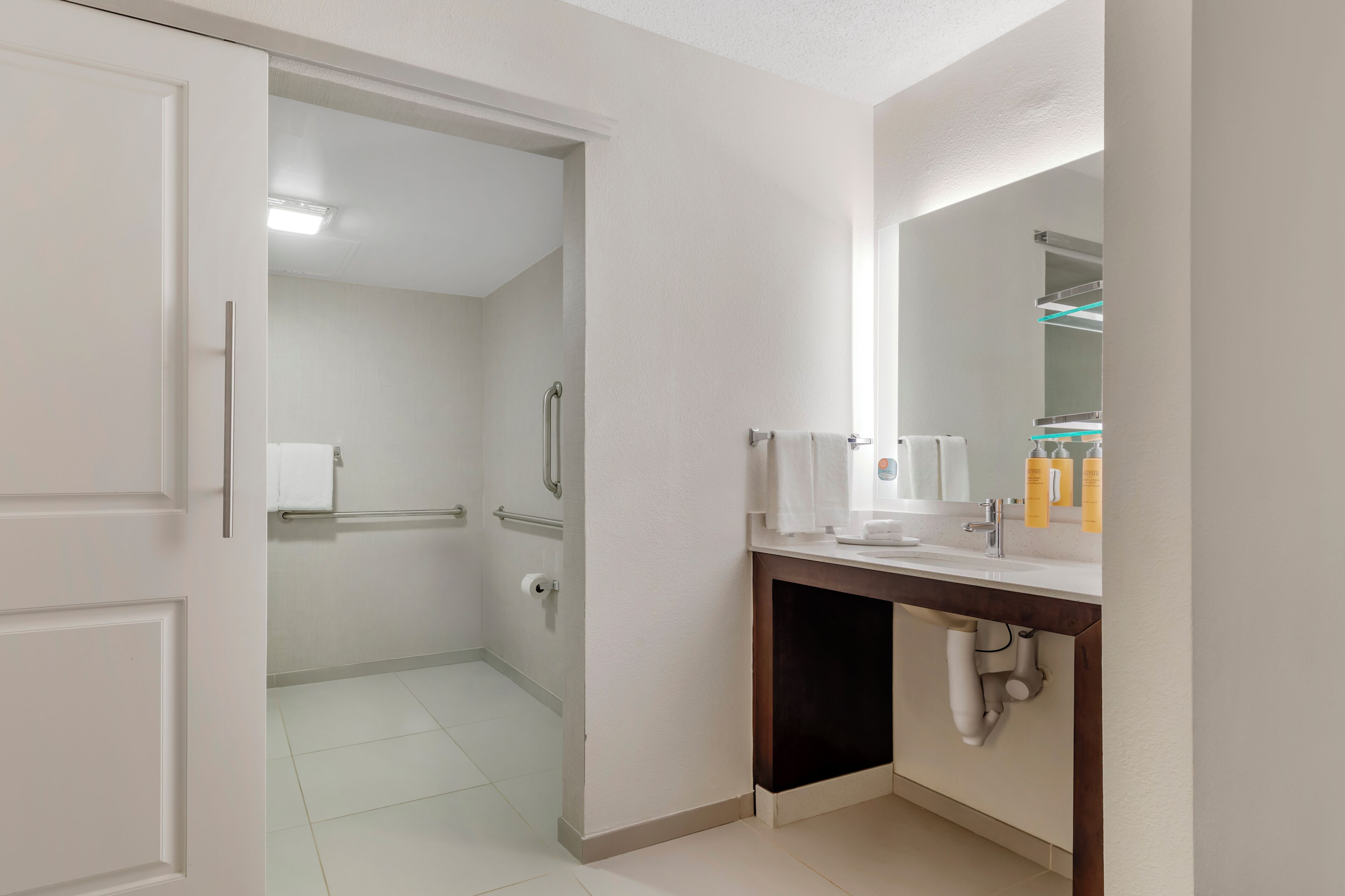 A modern bathroom with a toilet, sink, and mirror. There is a towel rack and a shower area visible in the background.